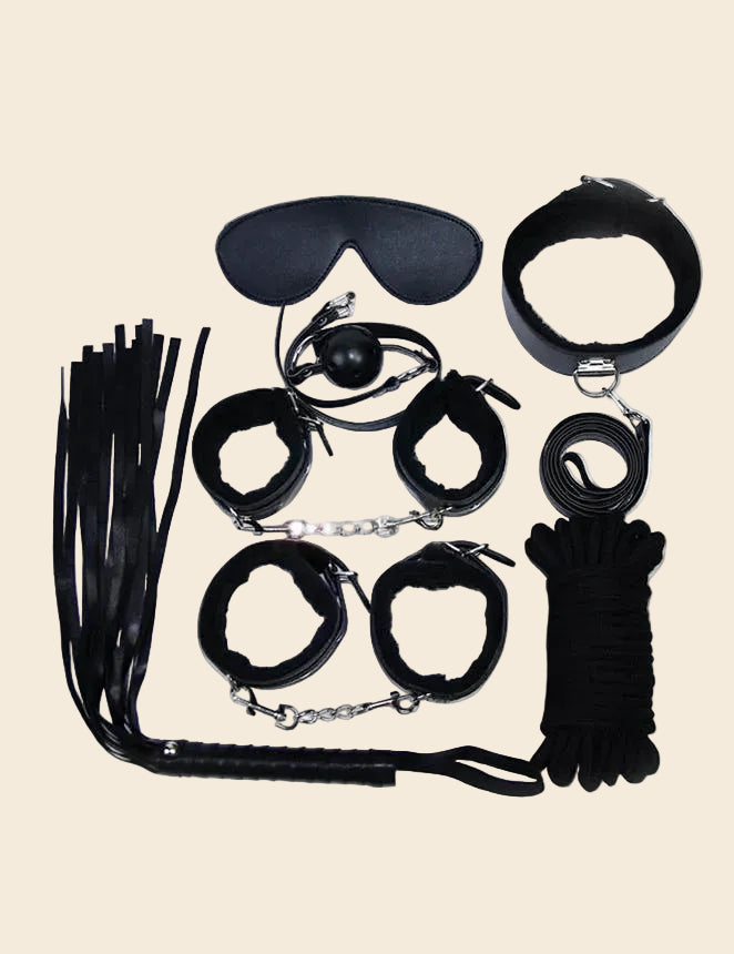A collection of black bondage accessories laid out on a white surface, including handcuffs, ankle cuffs, a collar with leash, a whip, a blindfold, and ropes.
